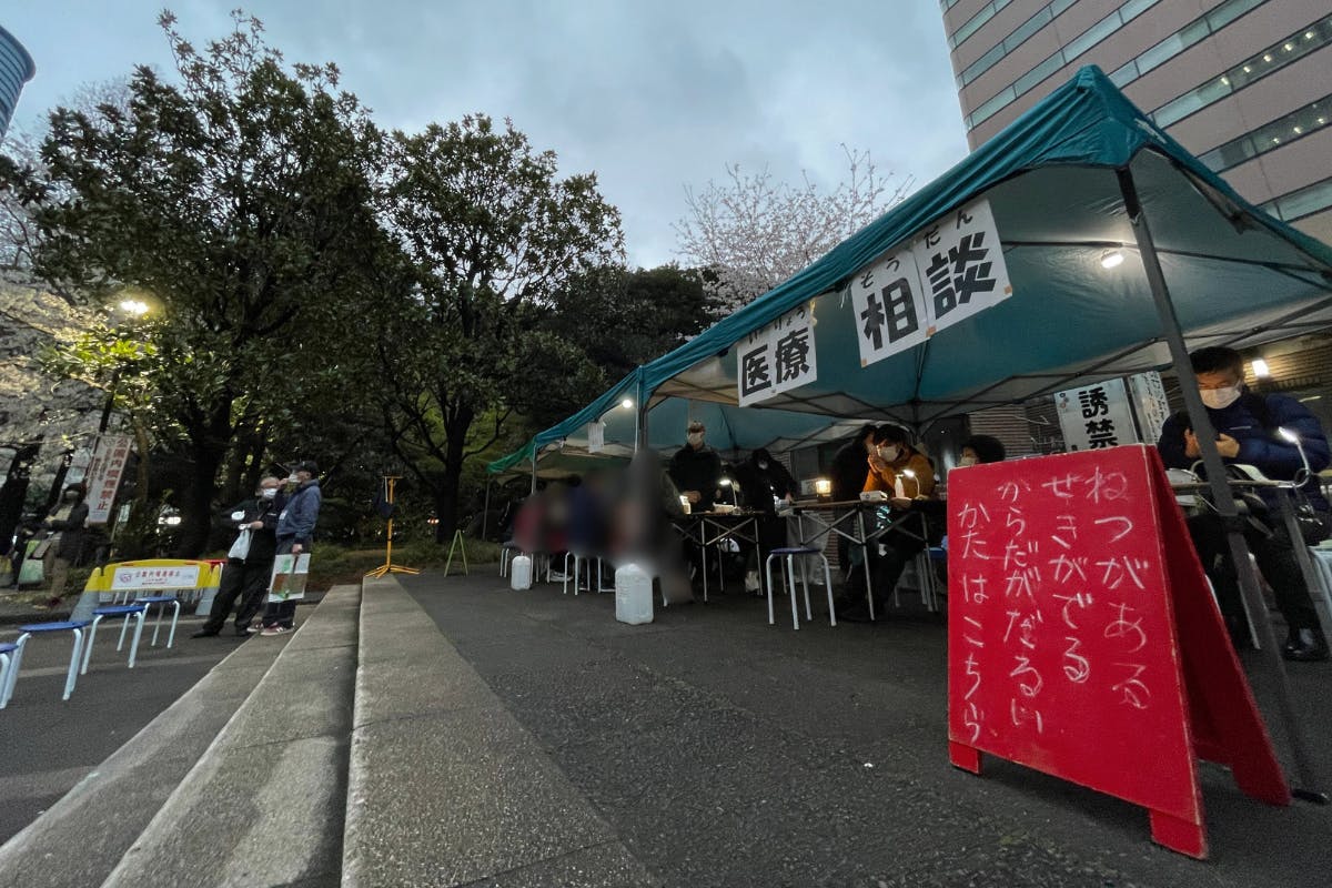 東京都内の公園での医療相談会の様子。悪天候の中、雨を避けるために、3張りのテントを立てて相談を受けている。新型コロナ感染症への感染を念頭に発熱者に対応するブースが手前に写っている。赤い看板に白いチョークで「ねつがある　せきがでる　からだがだるいかた　はこちら」と書かれている。