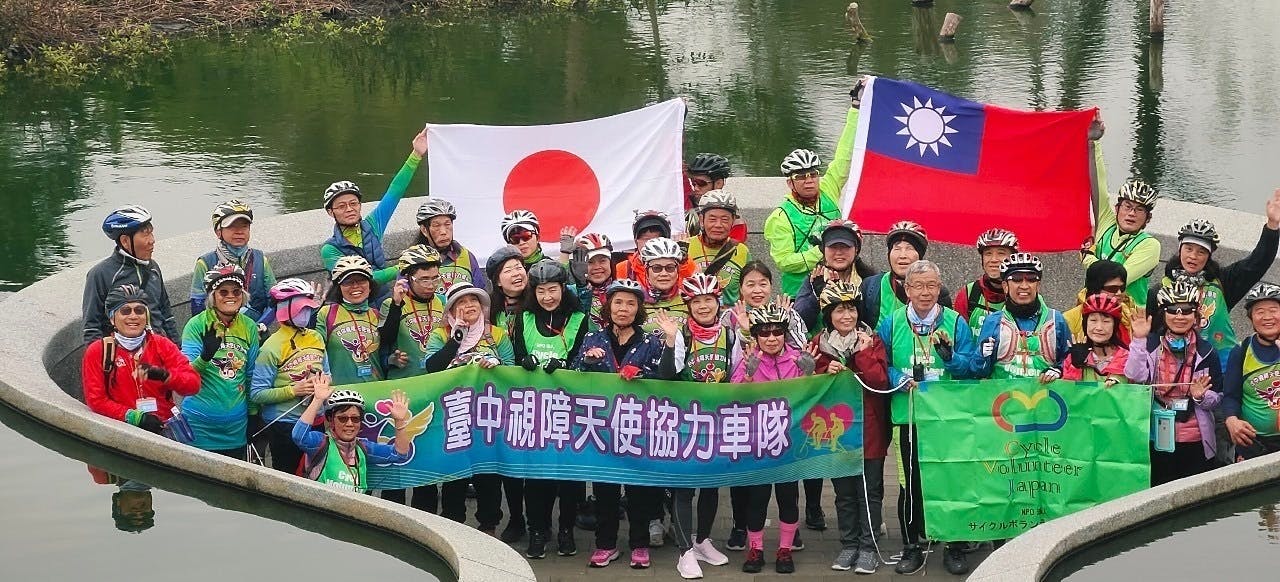 日本、台湾両国の国旗、自転車クラブの旗を持って50人くらいが川の中央部に立っている