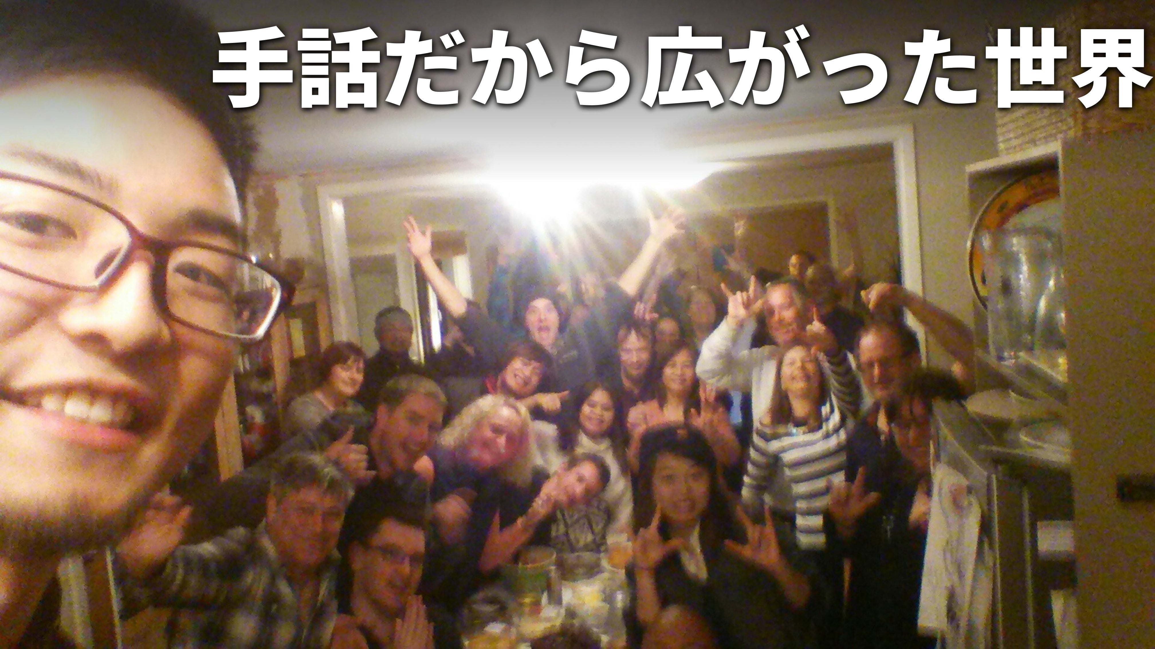 竹花がニュージーランドでホームパーティをした時に撮影した集合写真。老若男女の30人ほど写っている写真を背景に、白文字で「手話だから広がった世界」