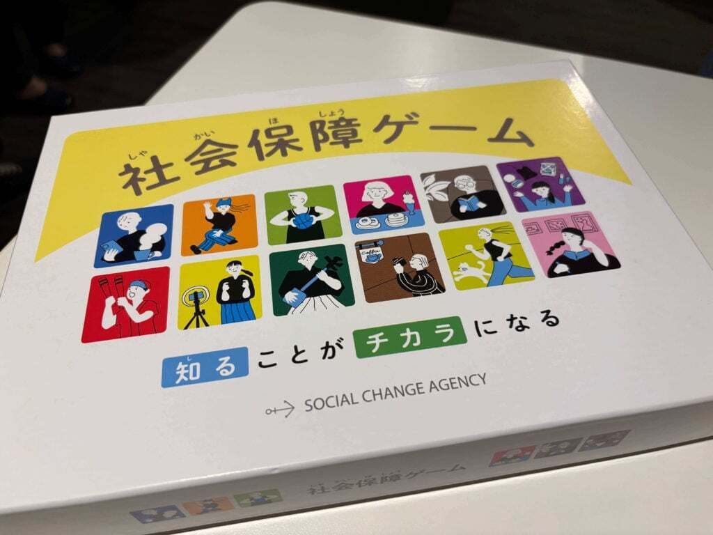 テーブルの上に置かれた『社会保障ゲーム』の箱。黄色と白のデザインで、表面には多様な人物が描かれたイラストが並んでいる。タイトルの下には『知ることがチカラになる』の文字と、SOCIAL CHANGE AGENCYのロゴが記されている。
