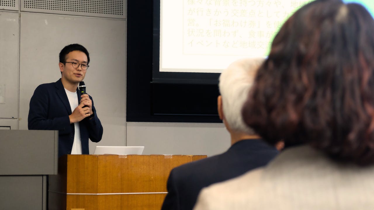 室内で行われている活動報告会の様子。眼鏡をかけ、紺色のジャケットを着たつくろい東京ファンド事務局長の大澤が木製の演台に立ち、マイクを両手で握って話しています。 大澤の背後にあるスクリーンにはスライド資料が投影されており、「様々な背景を持つ方々」「行きかう交差点」「『お福わけ券』」「地域交流」といった文字が一部読み取れます。手前には、話に耳を傾ける聴衆の後ろ姿が写っています。