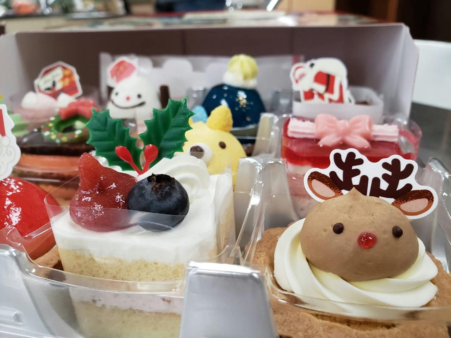 クリスマス仕様にデコレーションされた、色とりどりのミニケーキが箱に並んでいる接写写真。手前には、真っ白なホイップクリームに真っ赤なイチゴとブルーベリー、ヒイロゲンノウ（柊）の飾りが添えられた四角いケーキや、茶色のムースでトナカイの顔を模した丸いタルトが写っています。  奥には、黄色いクマの顔のケーキや、スノーマンやサンタクロースのピックが立てられたケーキが並び、パーティーの食卓を彩る賑やかで可愛らしい様子が伝わります。