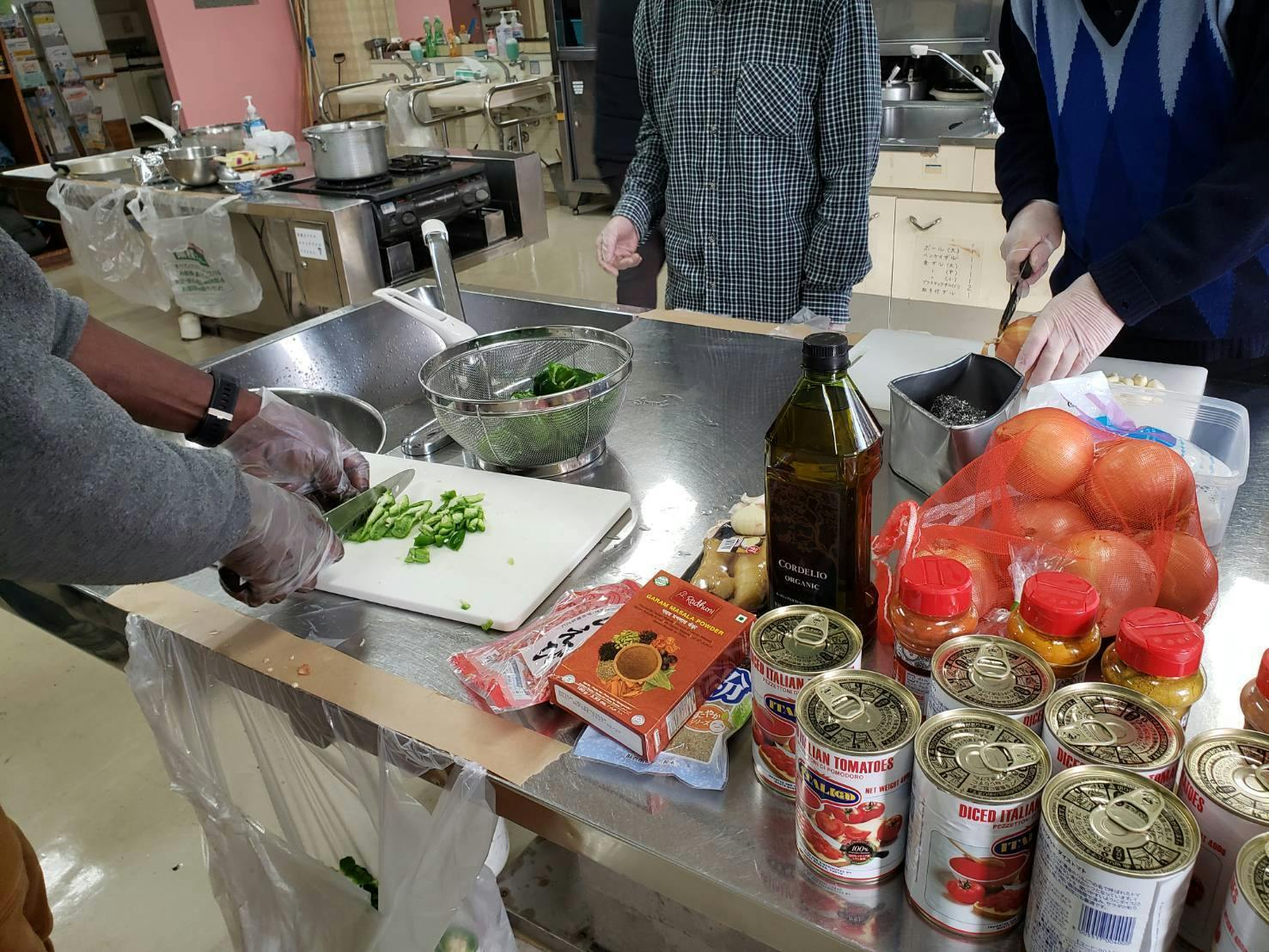 パーティーの料理を準備している厨房の様子。ステンレスの調理台を囲み、数人が手際よく作業しています。左手前ではビニール手袋をはめた人がピーマンを刻み、右手前では別の人が玉ねぎをスライスしています。  調理台の上には、大量のトマト缶、オリーブオイルの瓶、ガラムマサラなどのスパイスの箱、ネットに入ったたくさんの玉ねぎが所狭しと並んでおり、約40人分の多国籍な料理を総出で作っている活気ある現場の風景です。