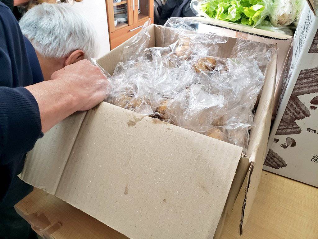 食料支援の梱包作業の様子。白髪のボランティアの方が、腰をかがめて大きな段ボール箱の中をのぞき込み、中身を整えています。  箱の中には、2〜3個ずつ小分けにされたジャガイモの袋が隙間なく並んでいます。