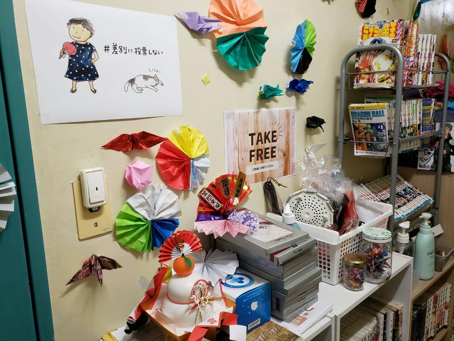 支援現場の壁際に飾られた、温かみのある年越しの風景。手前には、橙（だいだい）が乗った鏡餅と、「開運」「笑門来福」と書かれたお正月の縁起物が飾られています。  背景の壁には、色とりどりの折り紙で作られた扇や鶴が散りばめられ、「＃差別に投票しない」というメッセージが添えられた人物と猫のイラストポスターが貼られています。横には『ドラゴンボール』などの漫画が並ぶ本棚や「TAKE FREE」のコーナーもあり、誰もが自分らしく過ごせる居場所の日常と、新年を待つ静かな活気が伝わる一枚です。