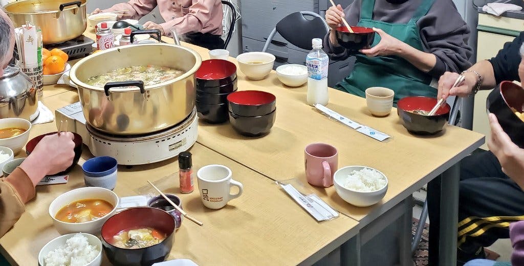 「鍋会(なべかい)」で、参加者が大きなテーブルを囲んで食事を楽しんでいる様子。 中央のテーブルには、カセットコンロに乗った大きな金の鍋が二つ置かれています。一つには大根おろしや野菜がたっぷりと入った「和風みぞれ鍋」が入っており、手前の器には赤みがかった「カレートマト鍋」が取り分けられています。 周囲には、白いご飯の入った茶碗、山盛りのみかん、マグカップなどが並び、エプロン姿のボランティアやシェルターの利用者、スタッフたちが箸を動かしながら共に食卓を囲んでいます。湯気の上がる温かい鍋を囲んで、国籍や立場を超えた人々が賑やかに交流している、冬の夜の温かな一場面です。