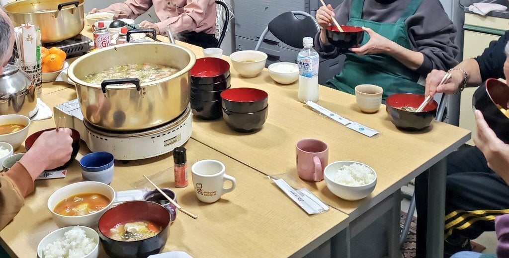 「鍋会（なべかい）」で、参加者が大きなテーブルを囲んで食事を楽しんでいる様子。  中央のテーブルには、カセットコンロに乗った大きな金の鍋が二つ置かれています。一つには大根おろしや野菜がたっぷりと入った「和風みぞれ鍋」が入っており、手前の器には赤みがかった「カレートマト鍋」が取り分けられています。  周囲には、白いご飯の入った茶碗、山盛りのみかん、マグカップなどが並び、エプロン姿のボランティアやシェルターの利用者、スタッフたちが箸を動かしながら共に食卓を囲んでいます。湯気の上がる温かい鍋を囲んで、国籍や立場を超えた人々が賑やかに交流している、冬の夜の温かな一場面です。