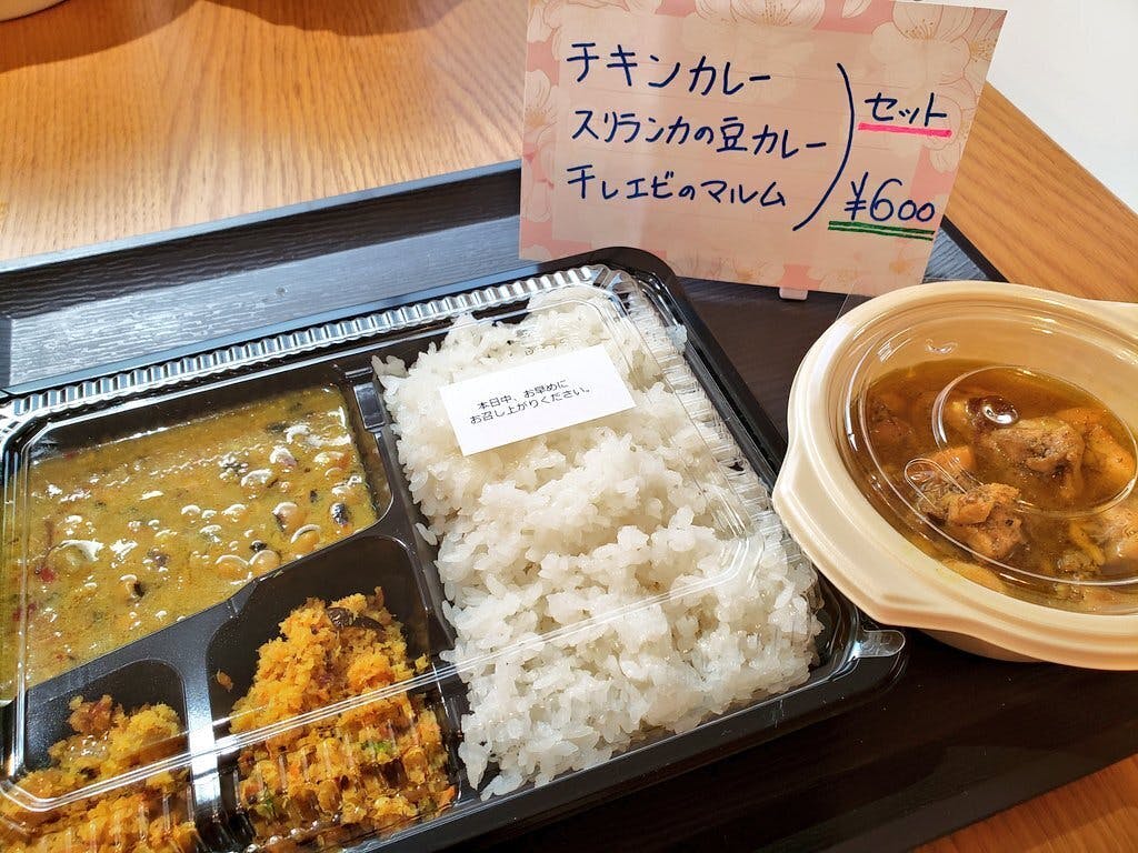 2月のチャリティーイベントで提供された「スリランカ・スペシャルセット」の実物写真。  黒いお弁当箱には、艶やかな白いご飯、スパイスの粒が見える黄色い豆カレー、そしてオレンジ色をした干しエビのマルム（和え物）がきれいに盛り付けられています。ご飯の上には「本日中、お早めにお召し上がりください」という小さな注意書きが添えられています。  隣には、ゴロゴロとした鶏肉が入ったチキンカレーの丸いカップが並んでいます。奥には手書きで「チキンカレー、スリランカの豆カレー、干しエビのマルム セット ￥600」と書かれた可愛らしい花のイラスト入りPOPが立てられており、オープン1時間で完売した活気あるイベントの様子を物語っています。