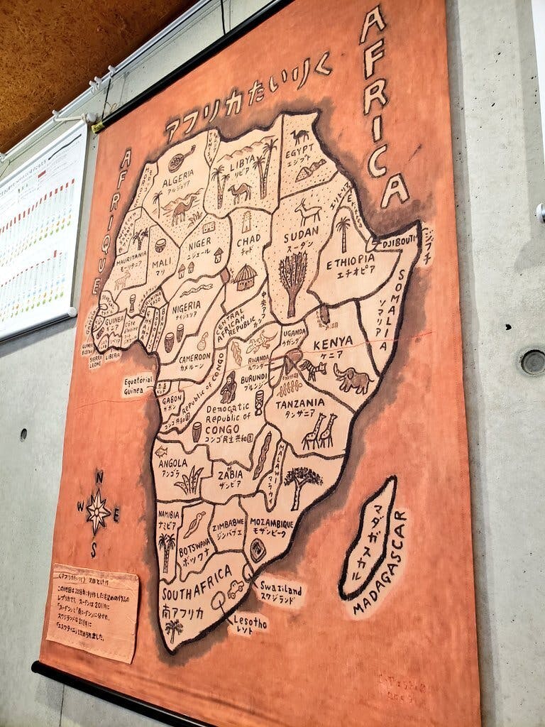 コンクリートの壁に掛けられた、オレンジ色の布に描かれた大きなアフリカ大陸の地図。  地図には「アフリカたいりく」と日本語で書かれ、各国の位置にアルジェリア、エジプト、ケニアなどの国名と、ラクダ、ゾウ、キリン、ワニといった動物や植物の可愛いイラストが手描きされています。  左下には、地図が作成された2008年以降の国名の変更（スーダンの分離やエスワティニへの改称など）を補足する手書きのメモが貼られています。西・中央アフリカ出身の方々が昔話を語り、歌い、踊ったというイベントの、文化的で豊かな国際交流の雰囲気を象徴する一枚です。