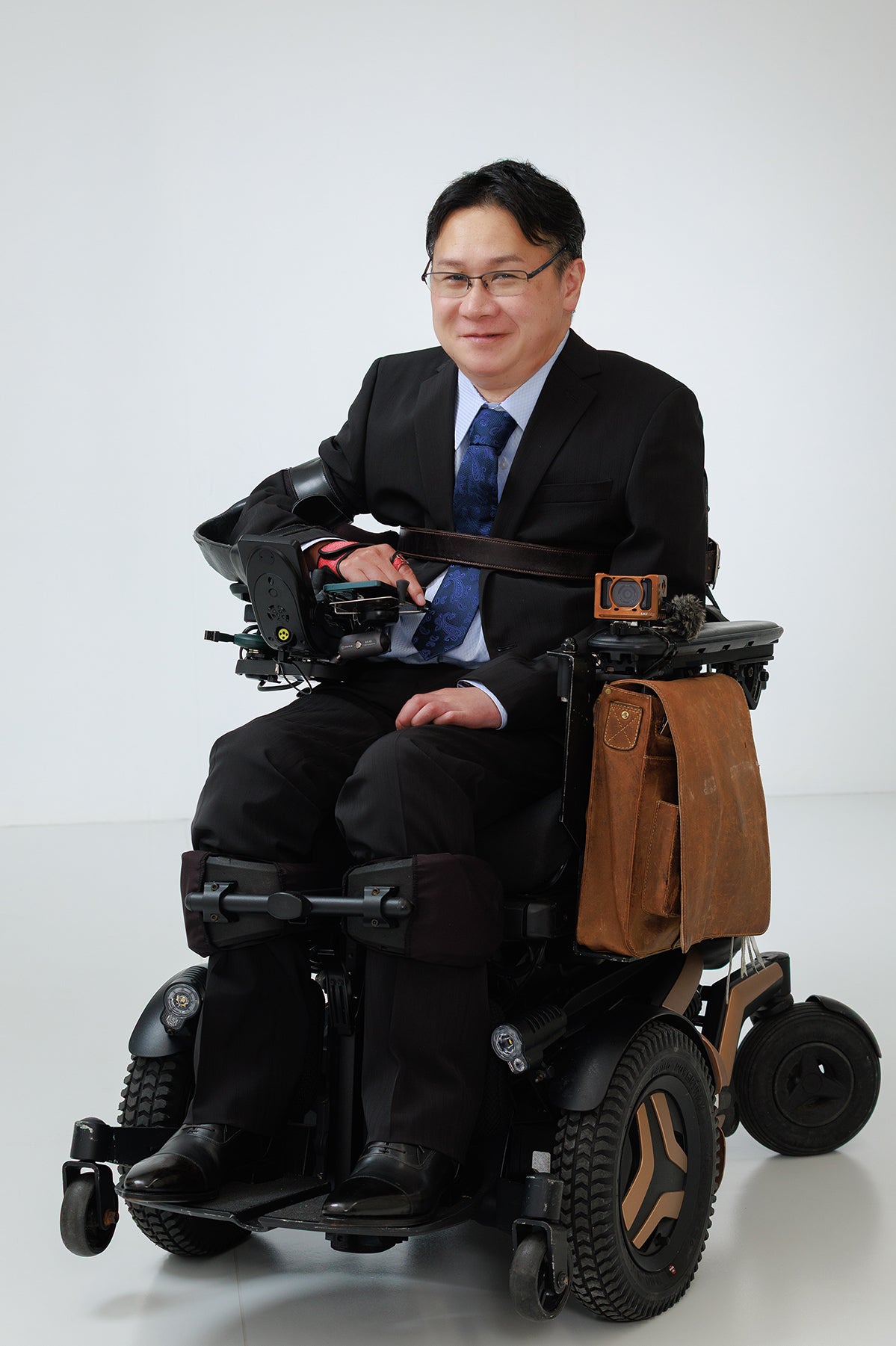 スーツを着て電動車椅子に乗り、微笑む眼鏡の男性の全身写真。カメラやバッグが装備された多機能な車椅子の様子。