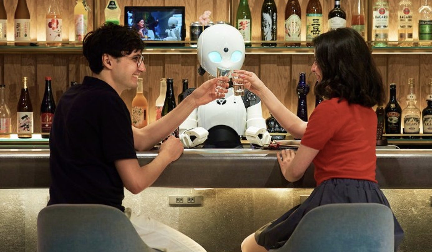 カウンターの中に立つ分身ロボットオリヒメDの前で笑顔で乾杯する男女の写真