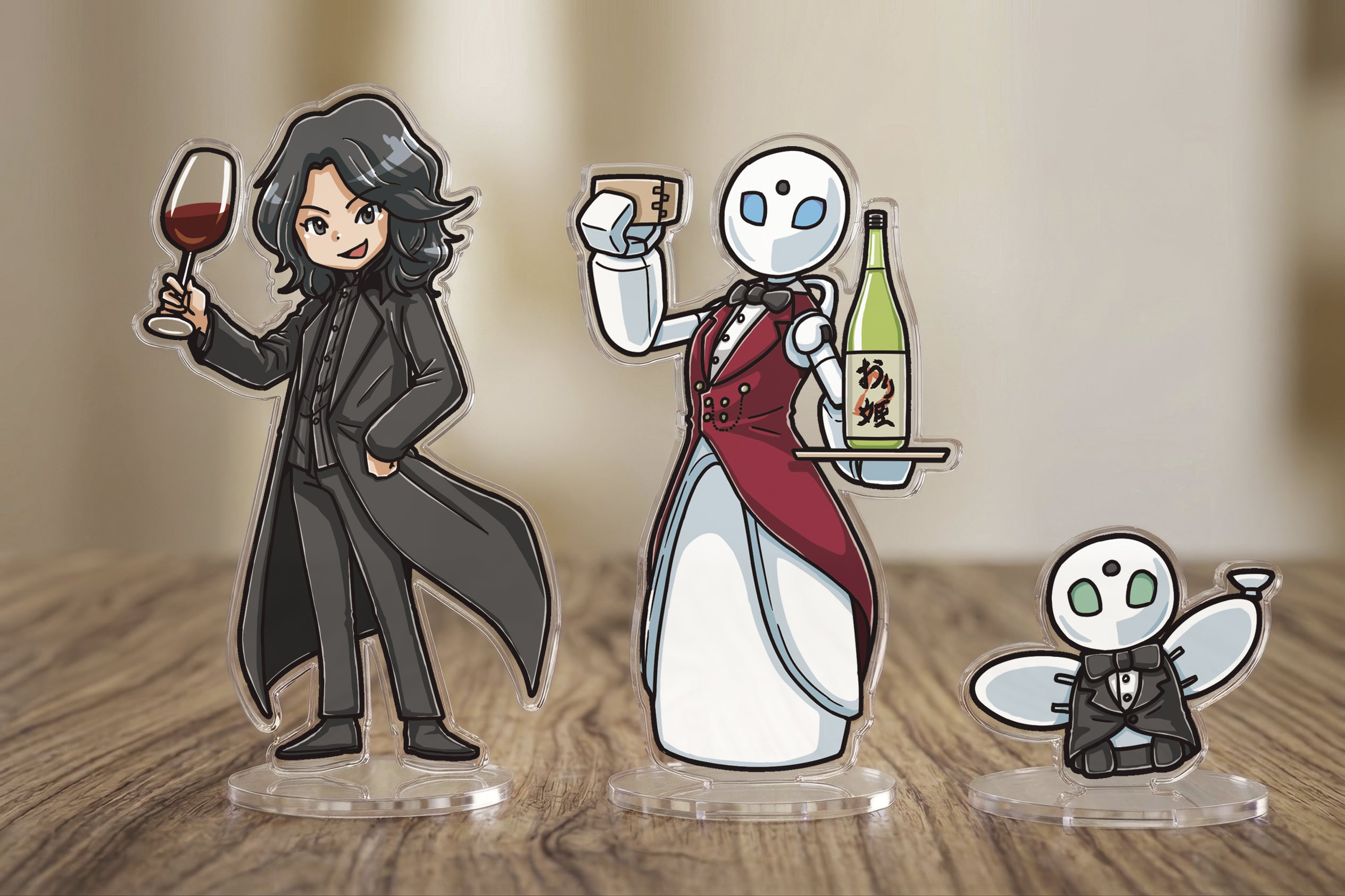 アクリルスタンド３点セットイメージ画像。左側は、吉藤オリィのキャラクター。トレードマークである黒い白衣を着て、左手に赤ワインの入ったグラスを持ち、右手をポケットに入れています。  中央は、分身ロボット「OriHime-D」。エンジ色のベストと蝶ネクタイ、白いロングスカートを着用しています。右手に枡（ます）を持ち、左手のトレイには「おり姫」と書かれた一升瓶を載せています。  右側は、小型の分身ロボット「OriHime」。黒いタキシードと蝶ネクタイを着用しています。左右の腕を挙げ、右手に小さな盃（さかずき）を持っています。
